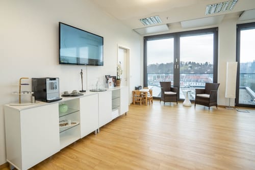 Wartebereich mit Kaffeestation, Bildschirm an der Wand und Sitzecke am Fenster